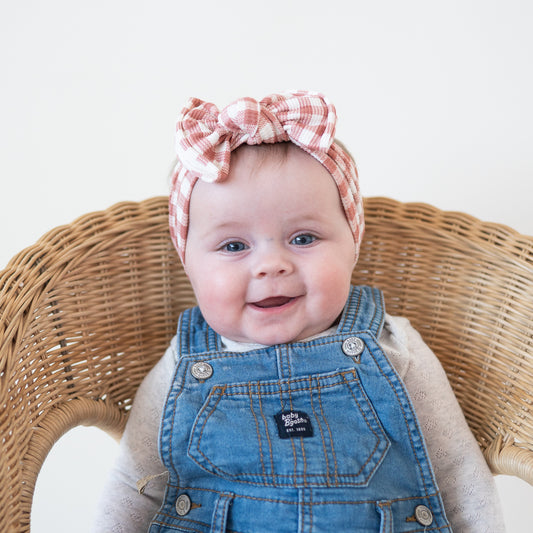 Baby wearing a checkered headband and denim overalls sitting in a wicker chair.