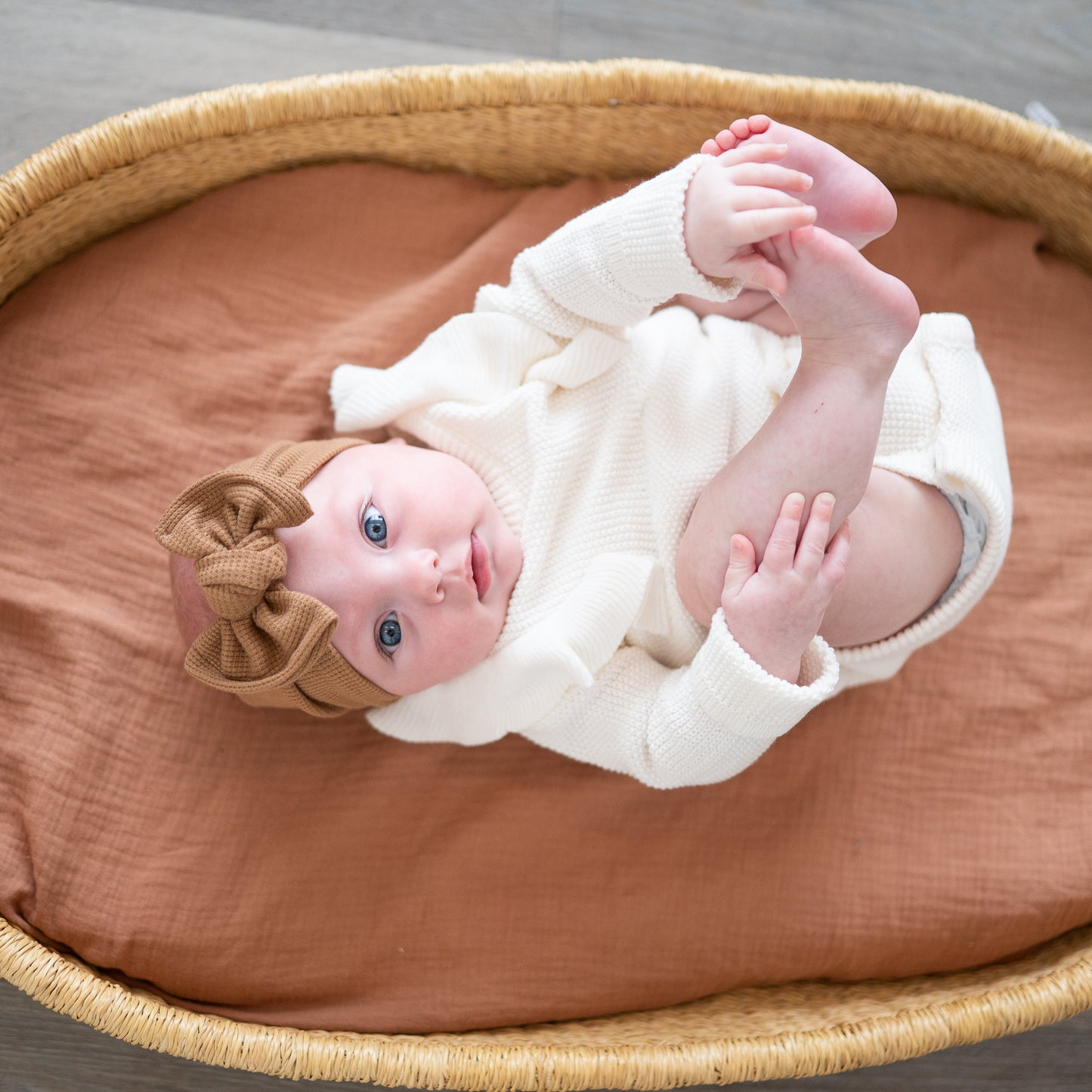 Baby in a white outfit with a brown bow lying in a Moses basket with a brown muslin blanket.