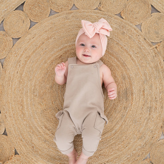 Baby in beige overalls and pink waffle oversized bow headband lying on a woven mat