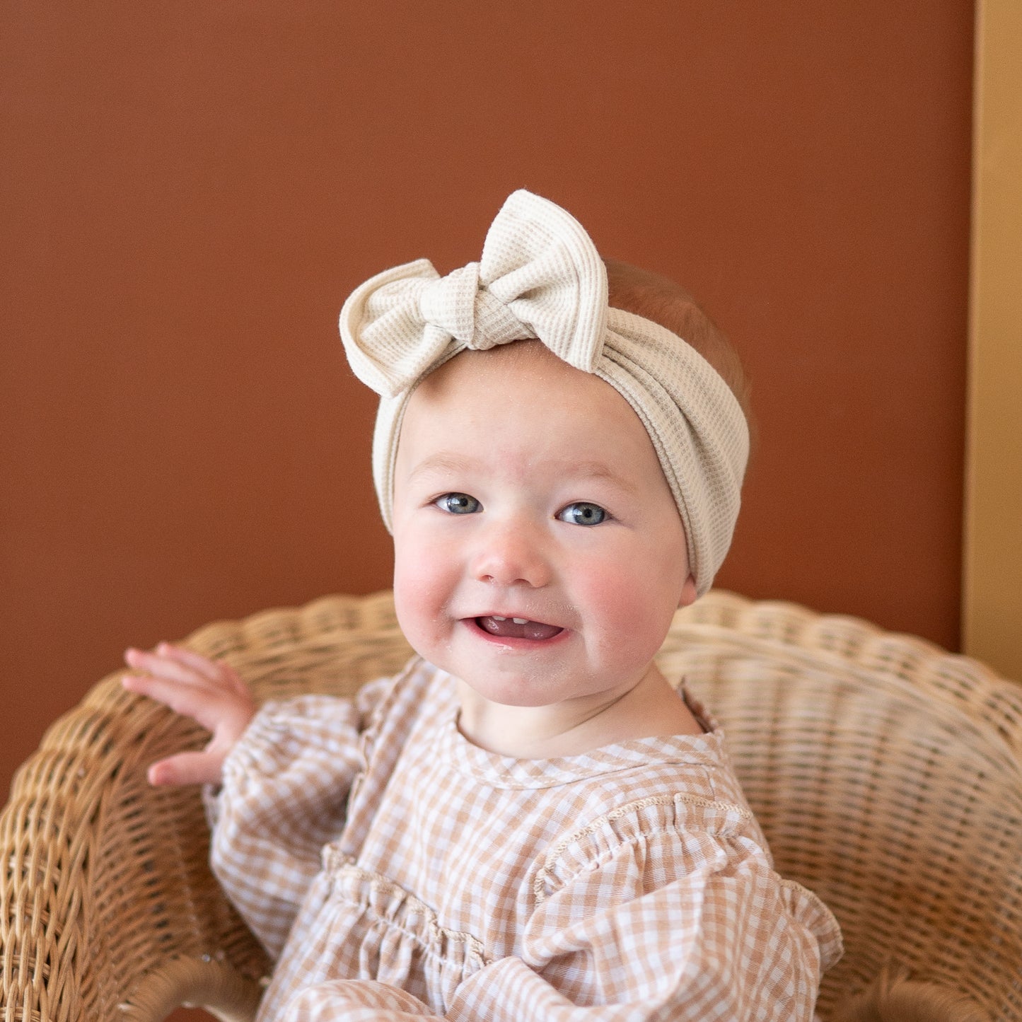 Baby wearing a beige headband with a bow, sitting in a wicker chair against a brown background