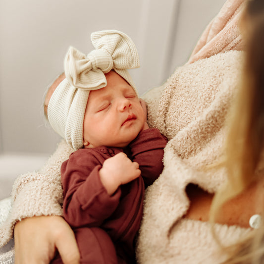 Newborn baby wearing a large beige waffle bow headband and brown outfit, held by an adult.