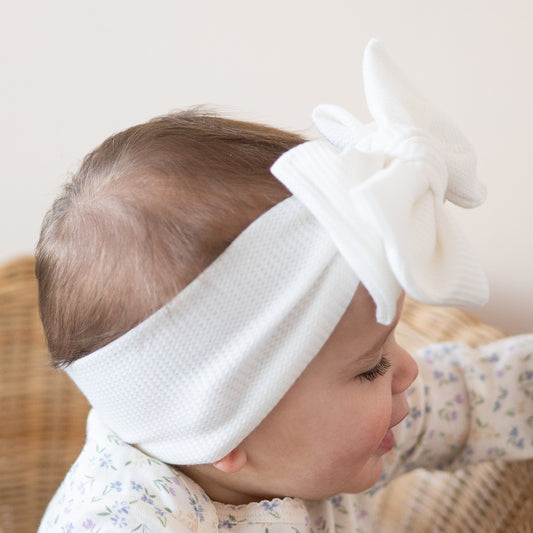 Side view of a baby wearing a white waffle oversized bow headband, sitting on a wooden surface.