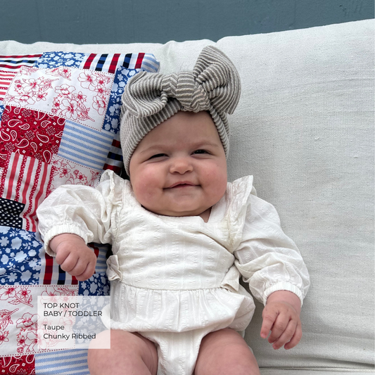 A baby wearing a chunky ribbed taupe top knot headband and a white long-sleeve romper, sitting on a white cushion next to a red, white, and blue patchwork pillow. A text overlay in the bottom left corner reads 'TOP KNOT BABY / TODDLER Taupe Chunky Ribbed'.