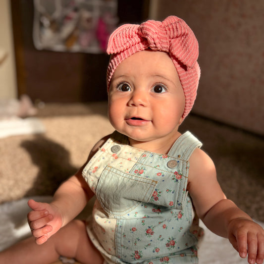 Baby wearing a pink headband and floral overalls sitting on a blanket.