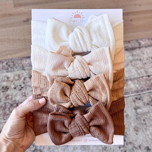 Set of pointelle knit textured baby bows in various neutral colors on a card held by a hand.
