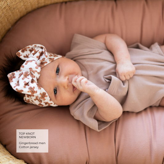 A newborn baby lying on a dusty rose cushion, wearing a taupe-brown ribbed cotton jersey romper and a white top knot headband with a brown gingerbread pattern. A text box in the bottom left corner reads 'TOP KNOT NEWBORN Gingerbread men Cotton jersey'.