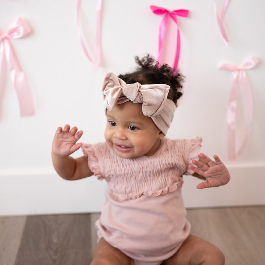 Baby wearing a pink outfit and headband with pink ribbons in the background