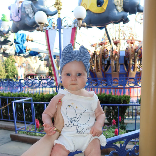 Baby in a Dumbo romper with Dumbo ride in the background wearing blue ribbed top knot.