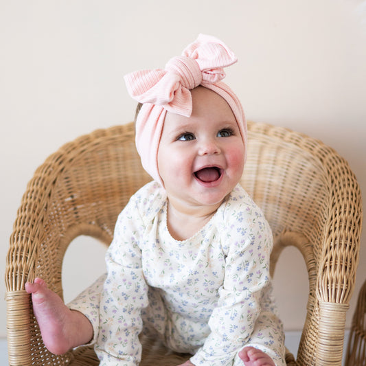Baby wearing a light pink ribbed headband with a large bow, sitting on a wicker chair.