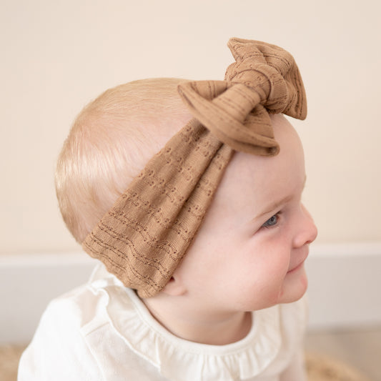 Side view of a baby wearing a mocha brown headband with a bow on a plain background.