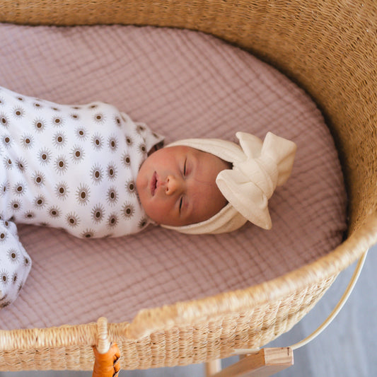 Newborn baby sleeping in a wicker crib with a white bow headband and patterned swaddle.