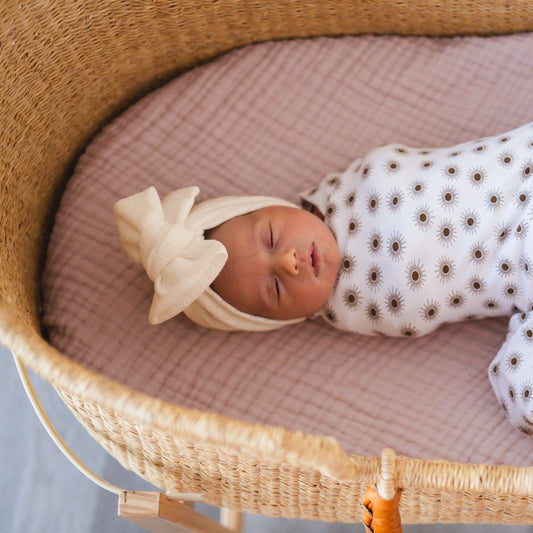 Newborn baby sleeping in a wicker crib with a white bow headband and patterned swaddle.