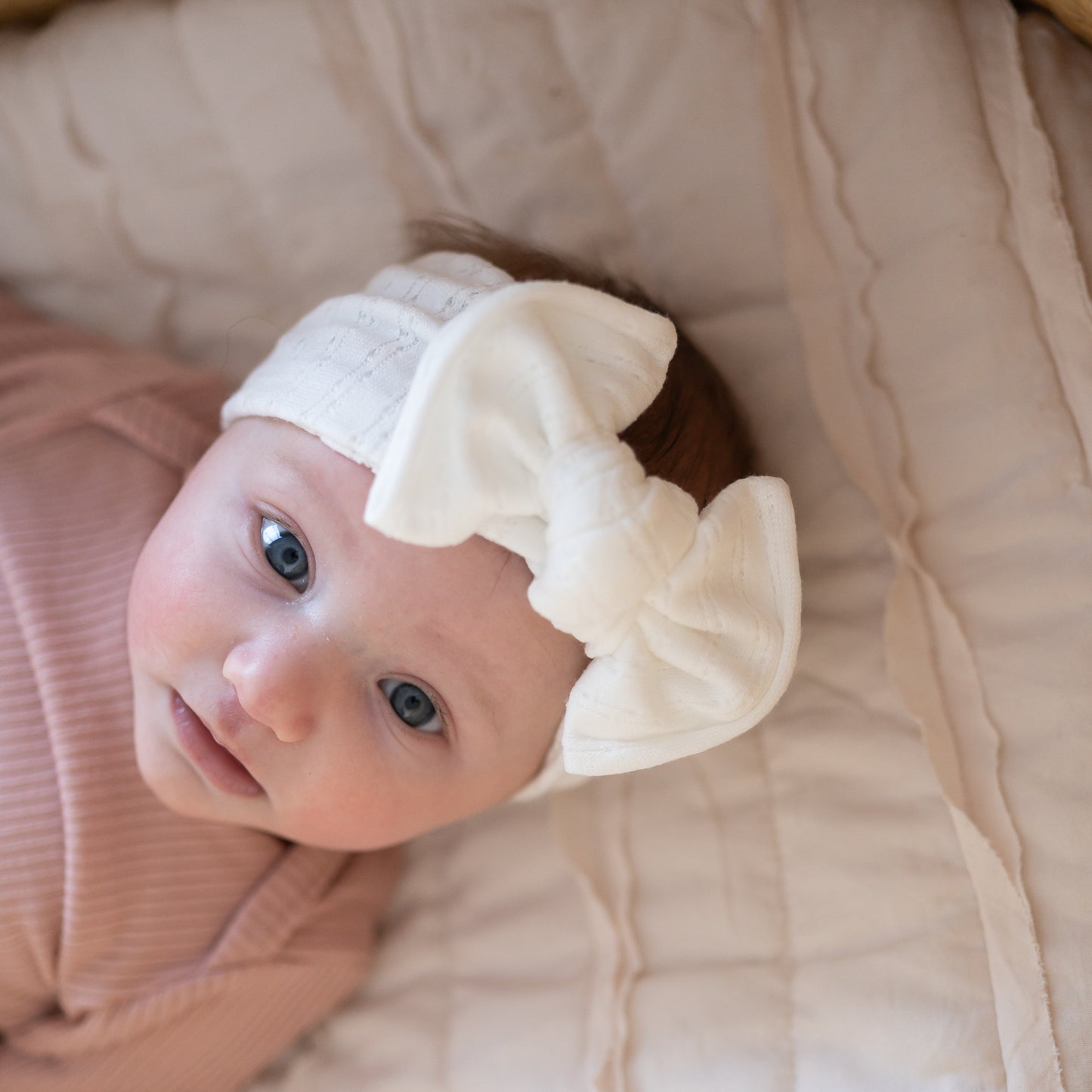 Baby wearing a white pointelle top knot headband with a bow, lying on a soft beige blanket.