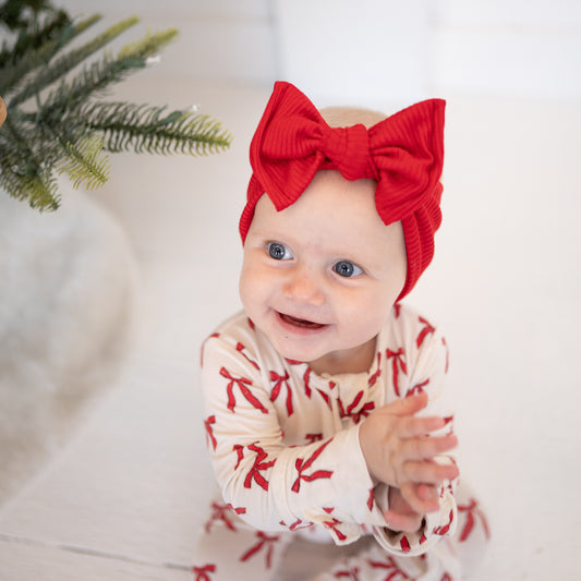 Baby wearing a red headband and white outfit with red patterns, sitting on a white surface.