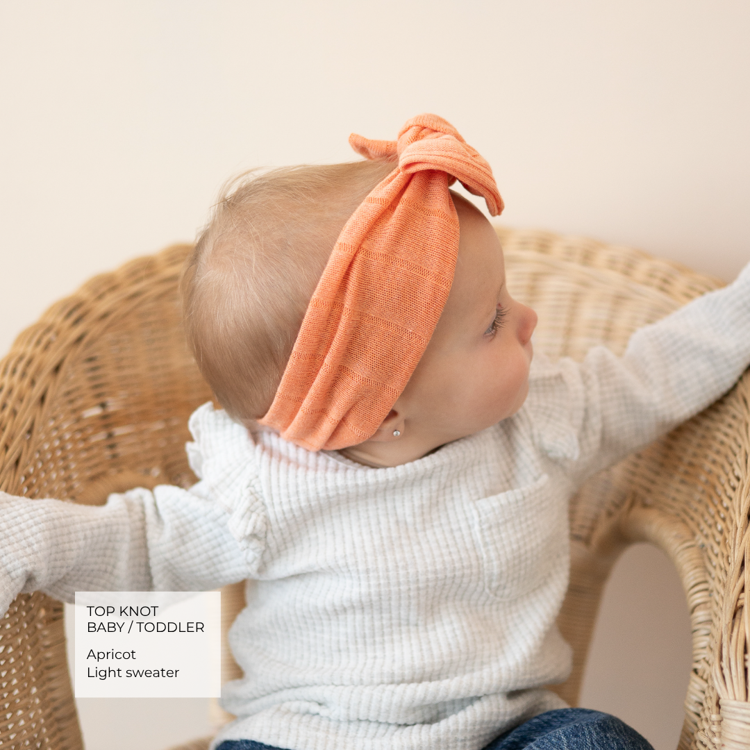 A baby wearing a light gray ribbed knit sweater and a peach apricot cotton headband with a top knot, sitting in a wicker chair. A text overlay in the bottom left reads 'TOP KNOT BABY / TODDLER Apricot Light sweater'.