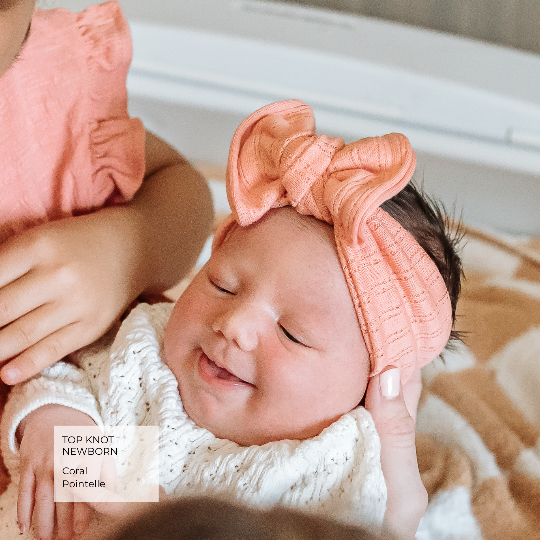 A newborn baby wearing a coral pink cotton top and matching bow headband, held by an adult in a soft indoor setting.