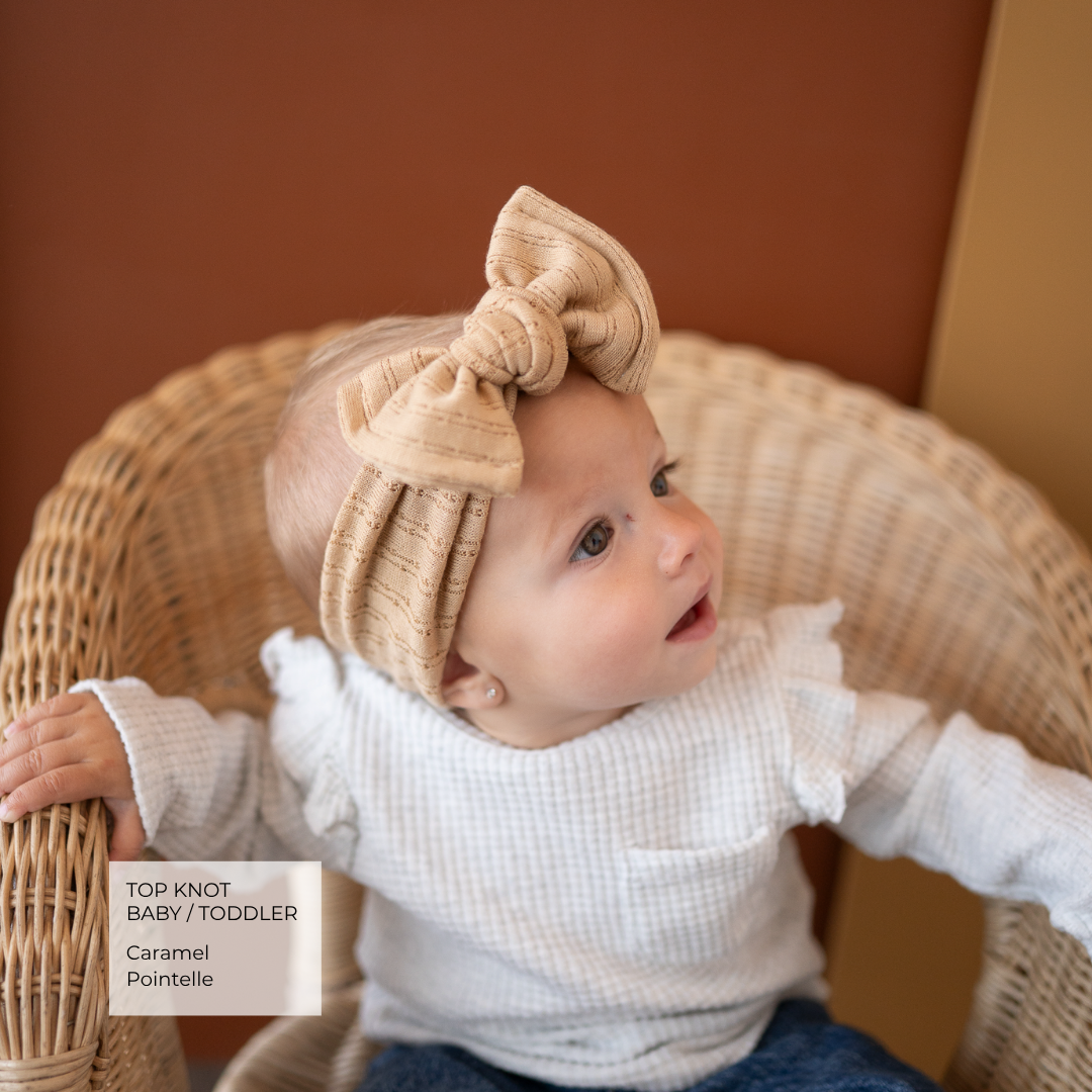 A baby wearing a beige top-knot bow headband and white textured long-sleeve top, sitting in a wicker chair against a warm brown wall. A text box in the lower left reads 'TOP KNOT BABY/TODDLER Caramel Pointelle'.
