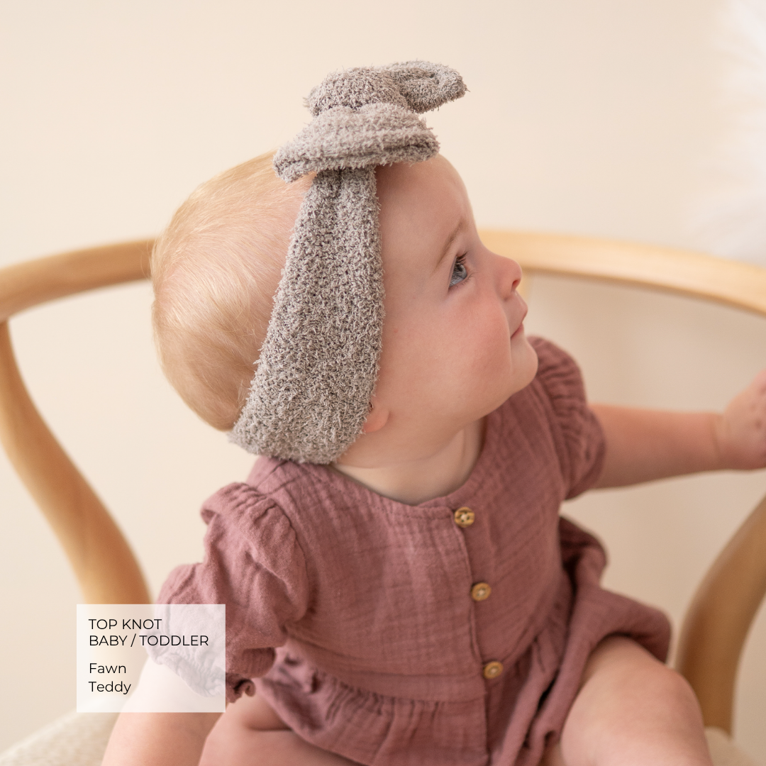 A baby wearing a textured gray top knot bunny-ear headband and a dusty rose linen romper, sitting in a wooden rocking chair. A text overlay in the bottom left reads 'TOP KNOT BABY / TODDLER Fawn Teddy'.