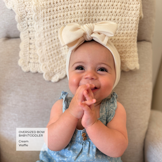A baby wearing an oversized cream bow baby/toddler headband and a light blue floral romper, sitting on a beige sofa with a crocheted cushion behind them. A text overlay in the bottom left reads 'OVERSIZED BOW BABY/TODDLER Cream Waffle'.