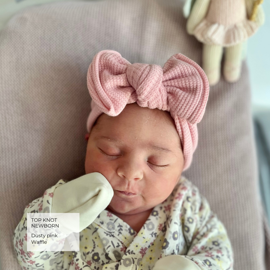 A newborn baby wearing a dusty pink knitted top knot headband with a large bow, lying on a soft beige blanket. The baby is dressed in a floral-patterned cream and pink outfit with matching cream booties.