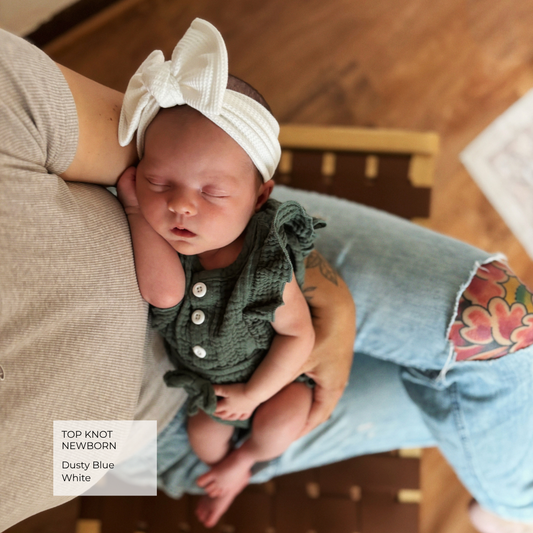 A newborn baby wearing a dusty blue knit romper and a white top knot headband, sleeping while being held by a person wearing light blue jeans with a floral patch on the knee.