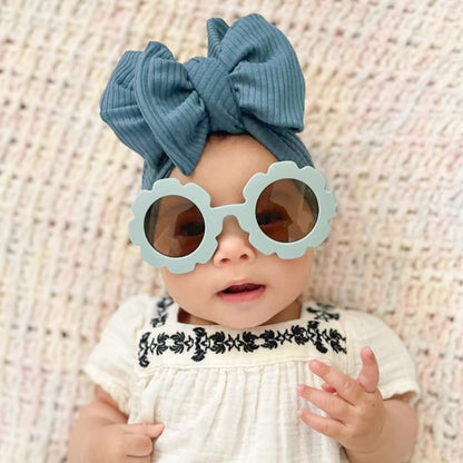 A baby wearing a blue ribbed headband with an oversized bow on top, in a white dress with black embroidery.