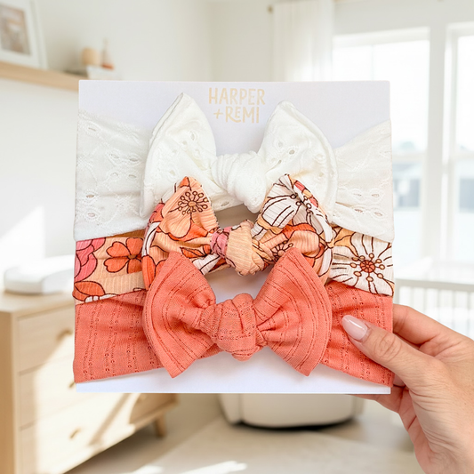 A baby girl's three-piece hair bow set by Harper + Remi, held by a hand against a bright, softly blurred nursery background. The set includes a white eyelet bow, a floral print bow in orange, peach, and cream, and a solid coral orange textured bow.