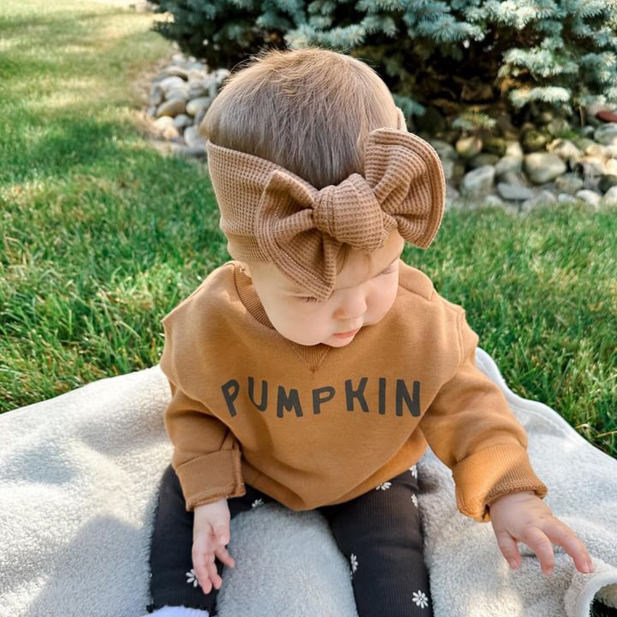 Baby wearing a 'PUMPKIN' sweatshirt and large bow headband outdoors.