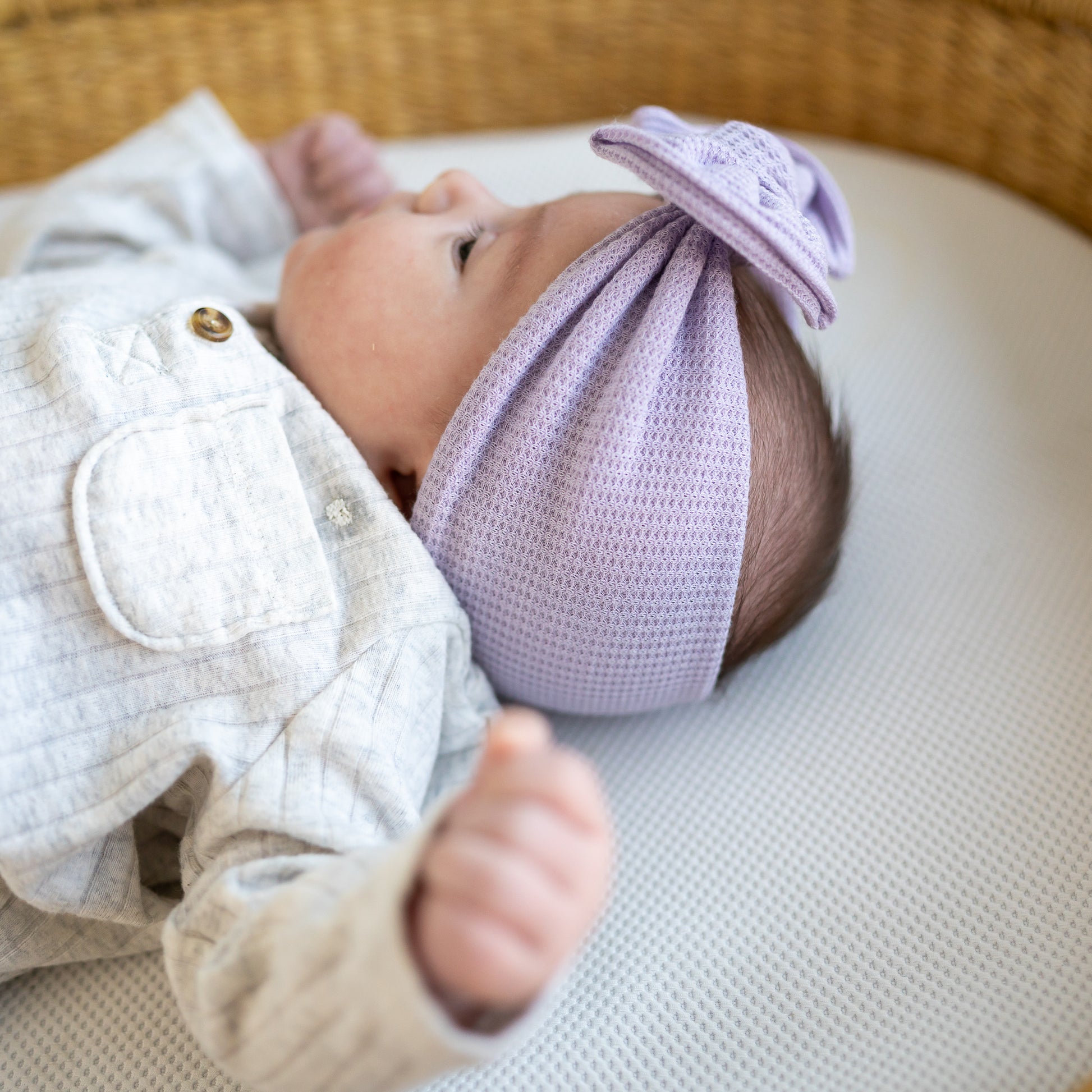 Newborn baby wearing a light purple waffle top knot headband lying on a textured surface