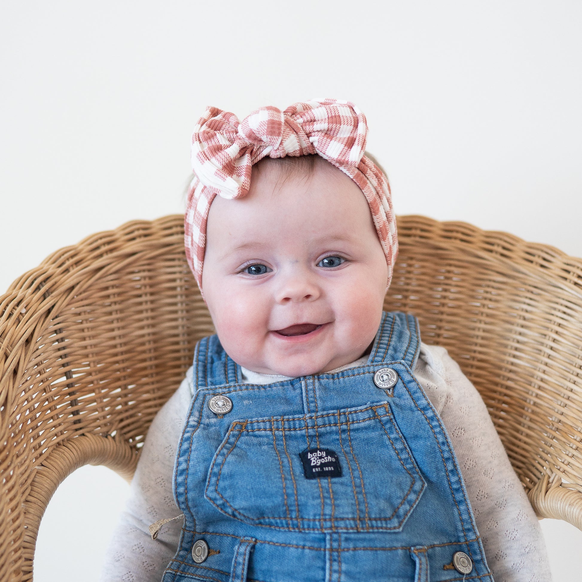 Baby wearing a checkered headband and denim overalls sitting in a wicker chair.