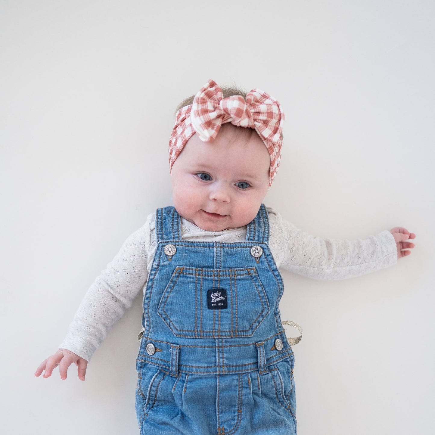 Baby wearing blue denim overalls and a pink gingham checkered headband on a white background