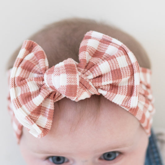 Detail view of a baby wearing a dusty pink and white checkered headband with a top knot on a plain background