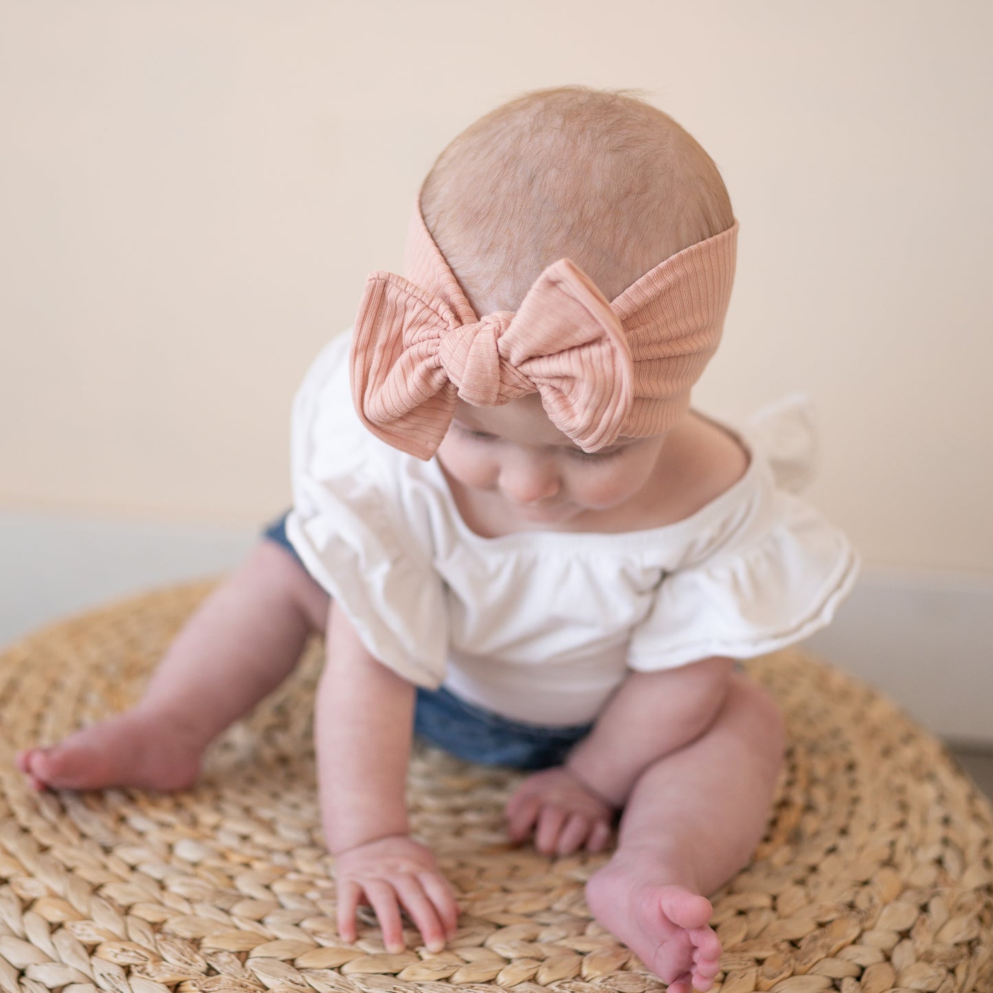 Baby sitting on a woven mat wearing a white outfit and dusty pink headband with a top knot bow.