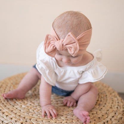 Baby sitting on a woven mat wearing a white outfit and dusty pink headband with a top knot bow.