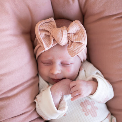 Newborn baby wearing a large bow headband on a dusty pink Snuggle Me Organic baby lounger.