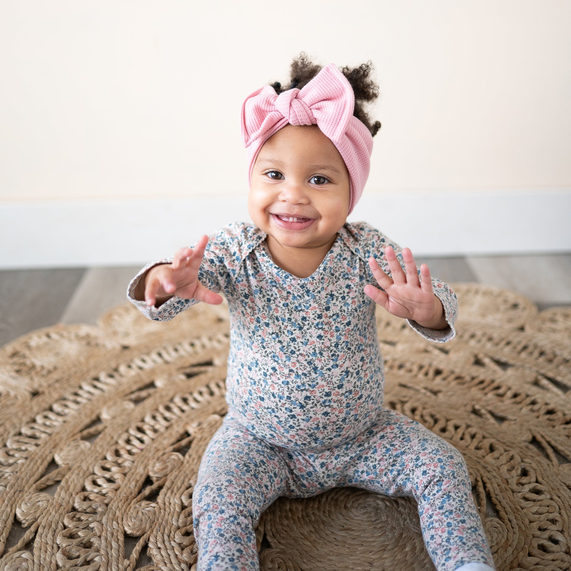 Baby wearing a floral onesie and pink headband sitting on a textured rug.