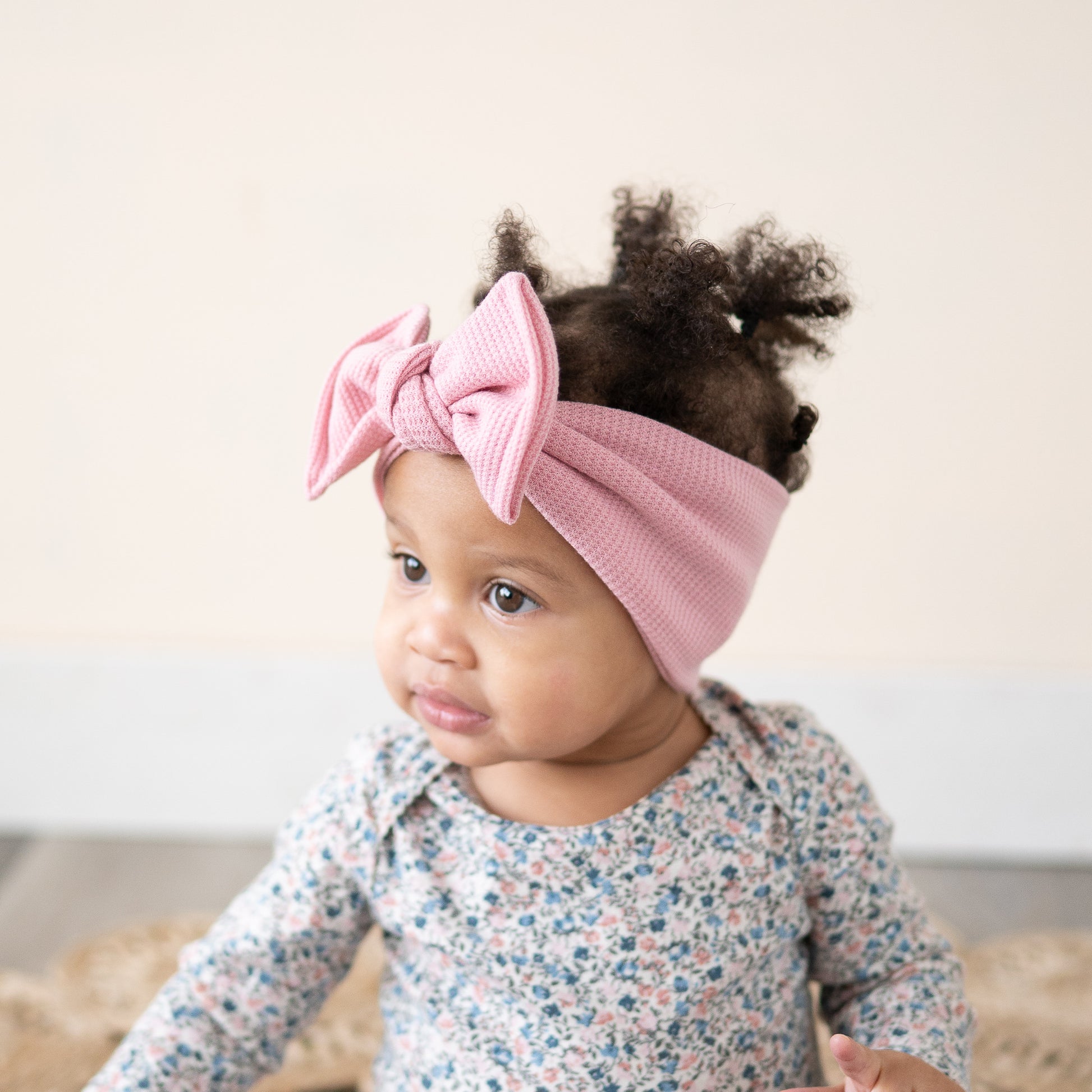 Baby wearing a pink headband with a bow, sitting on a woven rug.