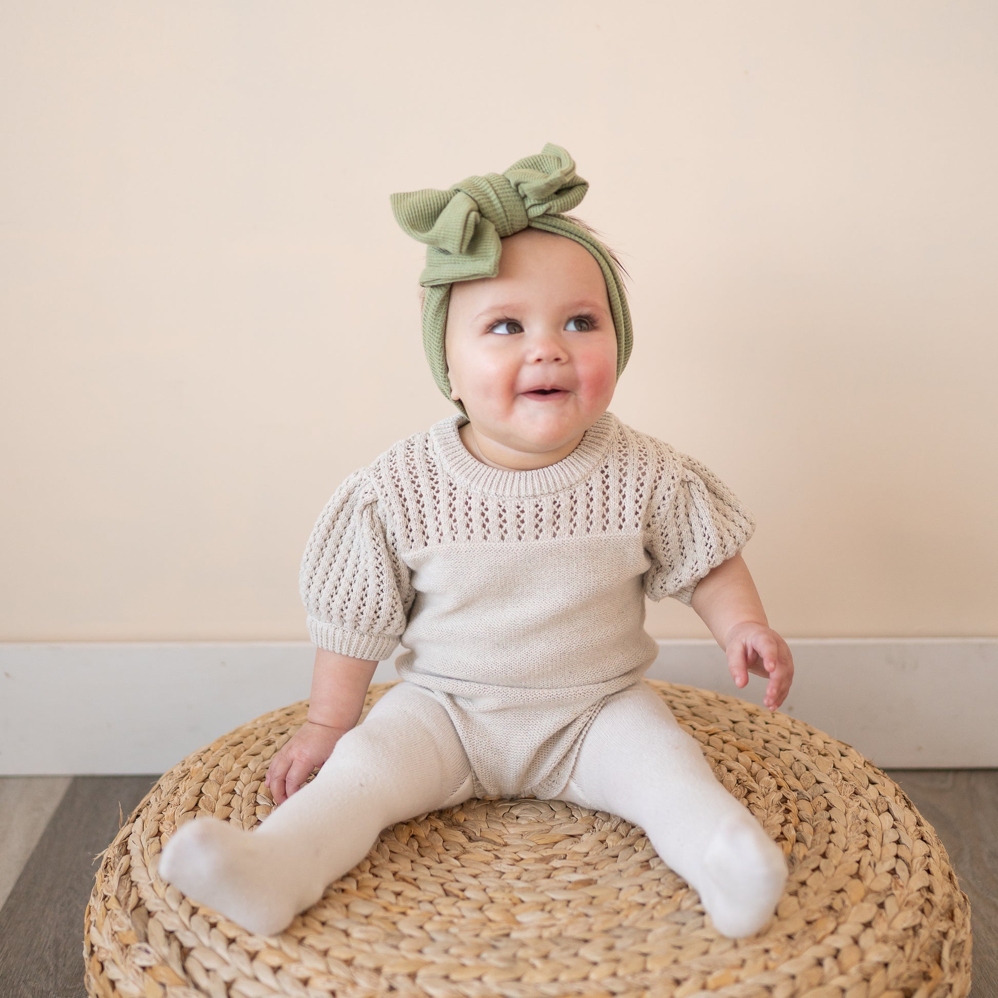 Baby sitting on a woven pouf wearing a knitted outfit and green headband.