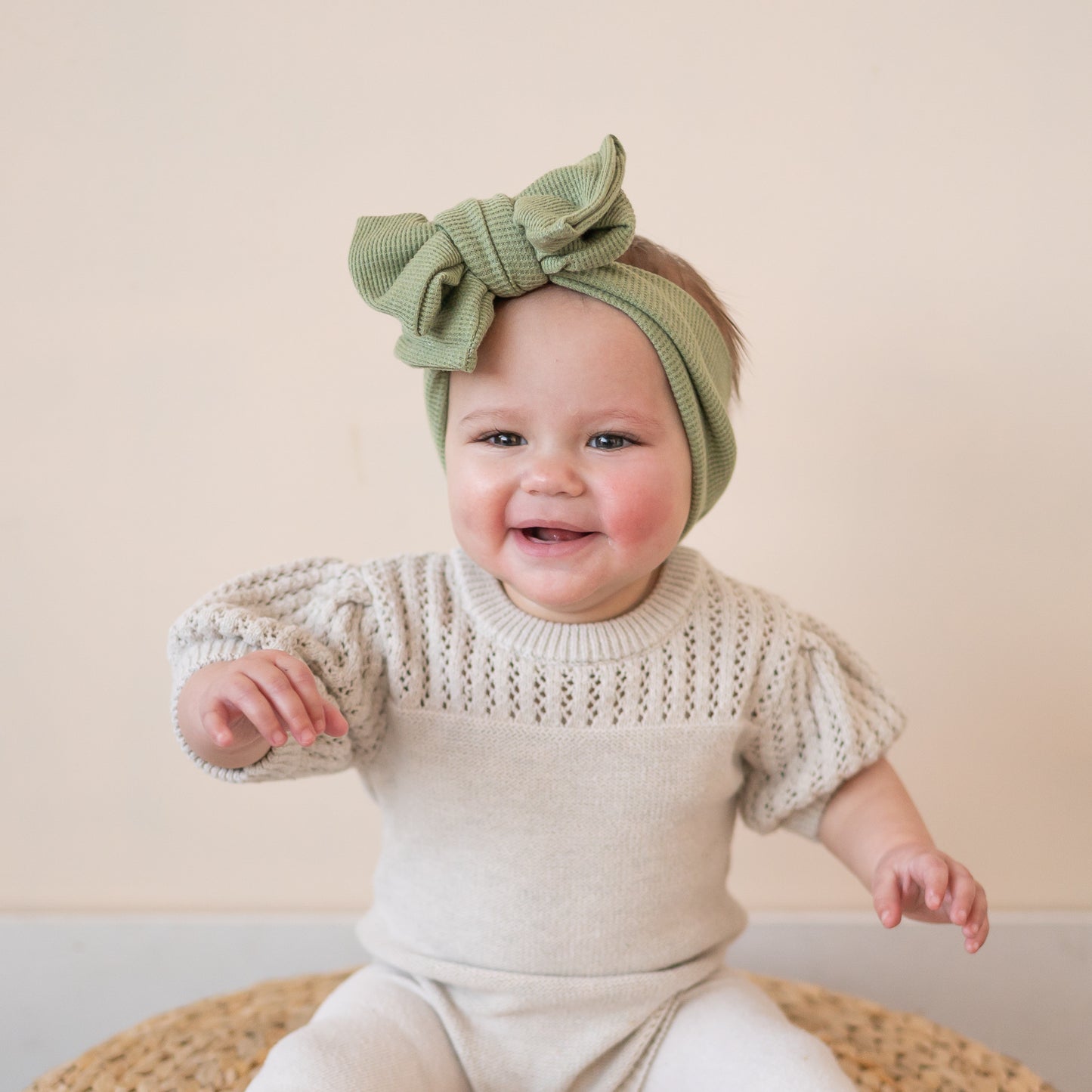 Baby wearing a green oversized bow headband and white outfit against a beige background