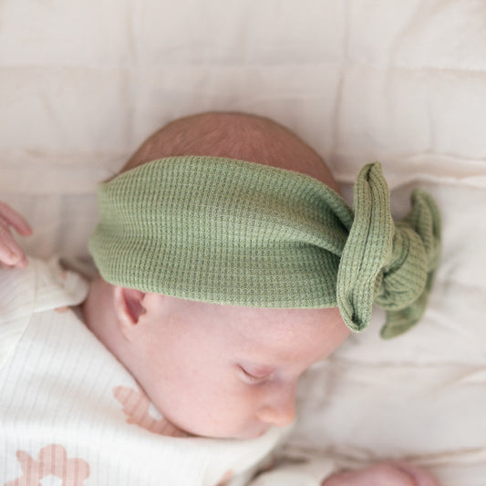 Side view of a newborn baby wearing a green knotted headband on a soft white background