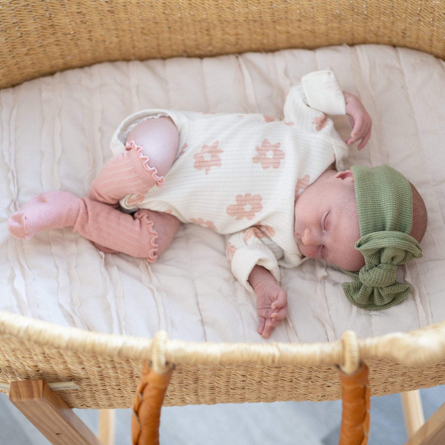 Newborn baby sleeping in a Moses basket wearing a floral onesie and green waffle top knot headband.