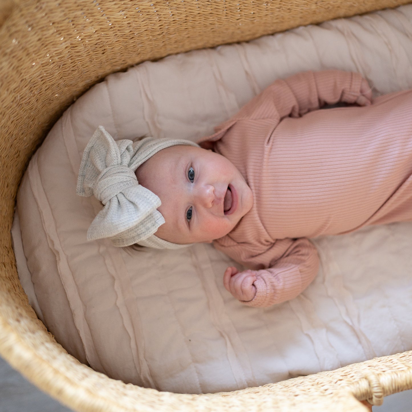 Newborn baby in a pink outfit with a heather gray waffle oversized bow headband lying in a woven basket.