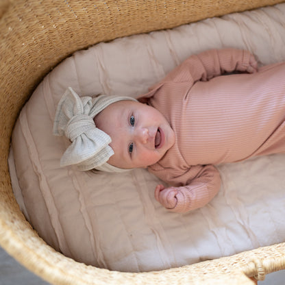 Newborn baby in a pink outfit with a heather gray waffle oversized bow headband lying in a woven basket.