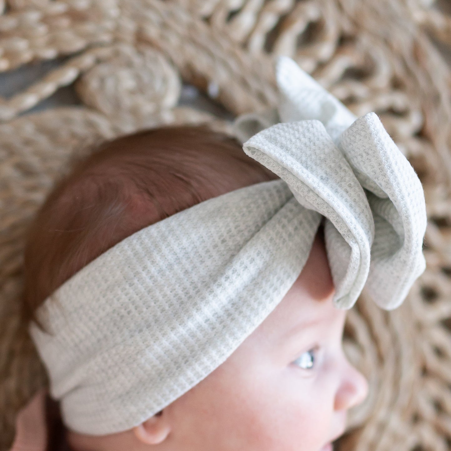 Baby wearing a light gray headband with a bow, lying on a woven surface.