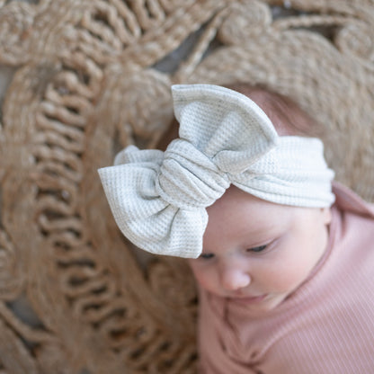 Baby wearing a large light gray oversized bow headband with a textured wall in the background
