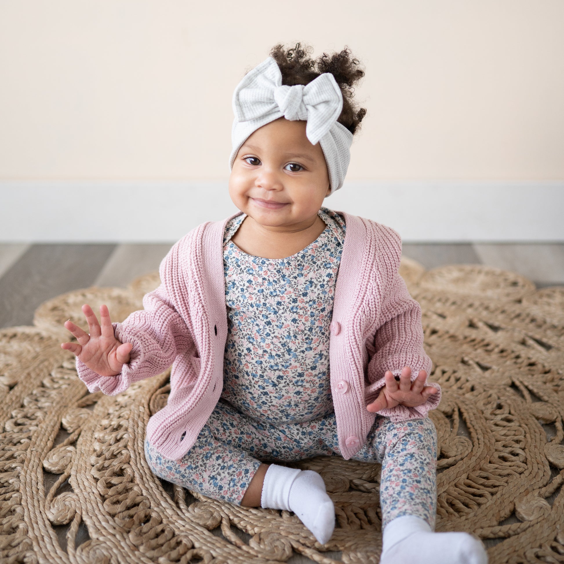 Baby girl wearing a gray waffle top knot, pink cardigan and floral outfit sitting on a textured rug.