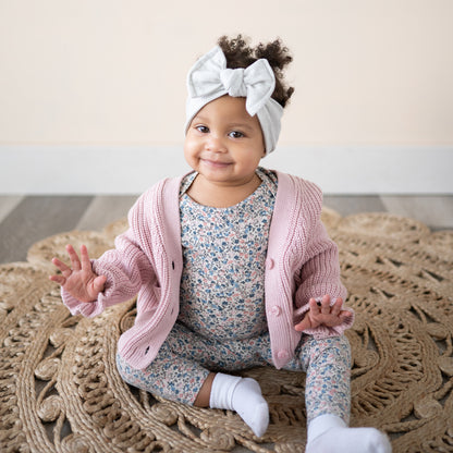 Baby girl wearing a gray waffle top knot, pink cardigan and floral outfit sitting on a textured rug.