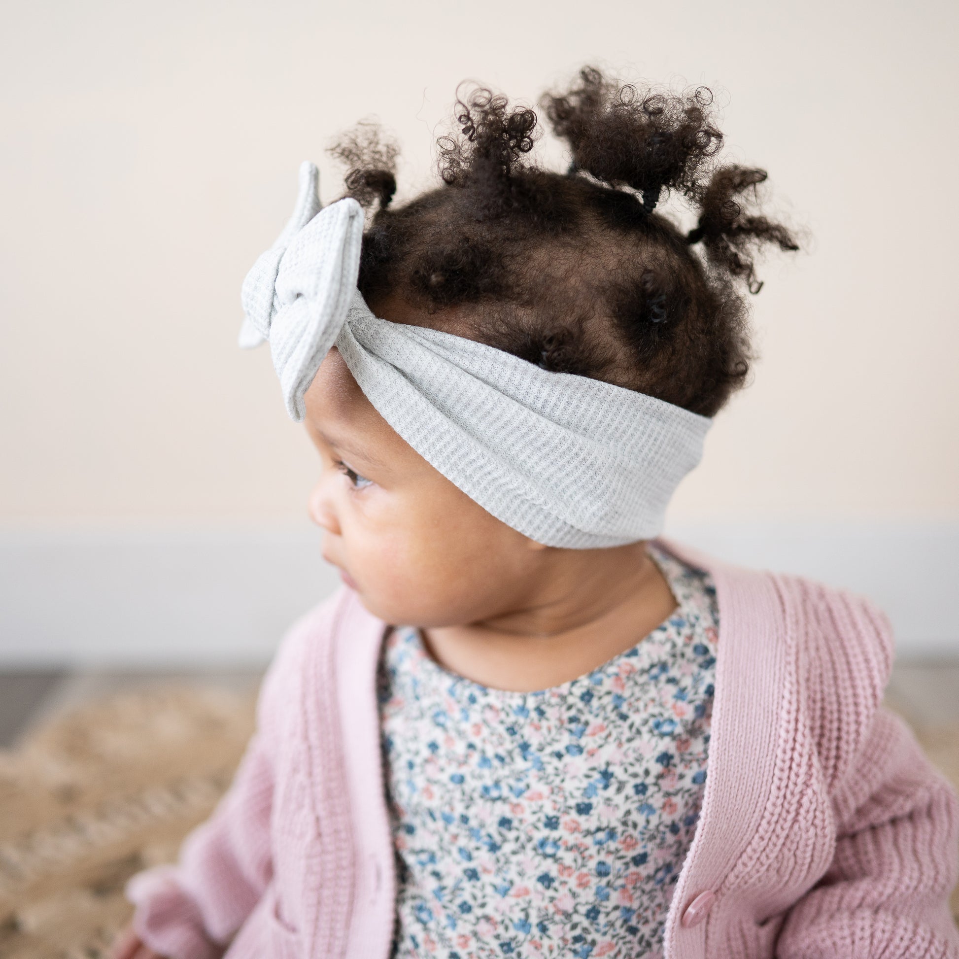 Baby girl wearing a light gray headband with a bow, pink cardigan, and floral shirt.