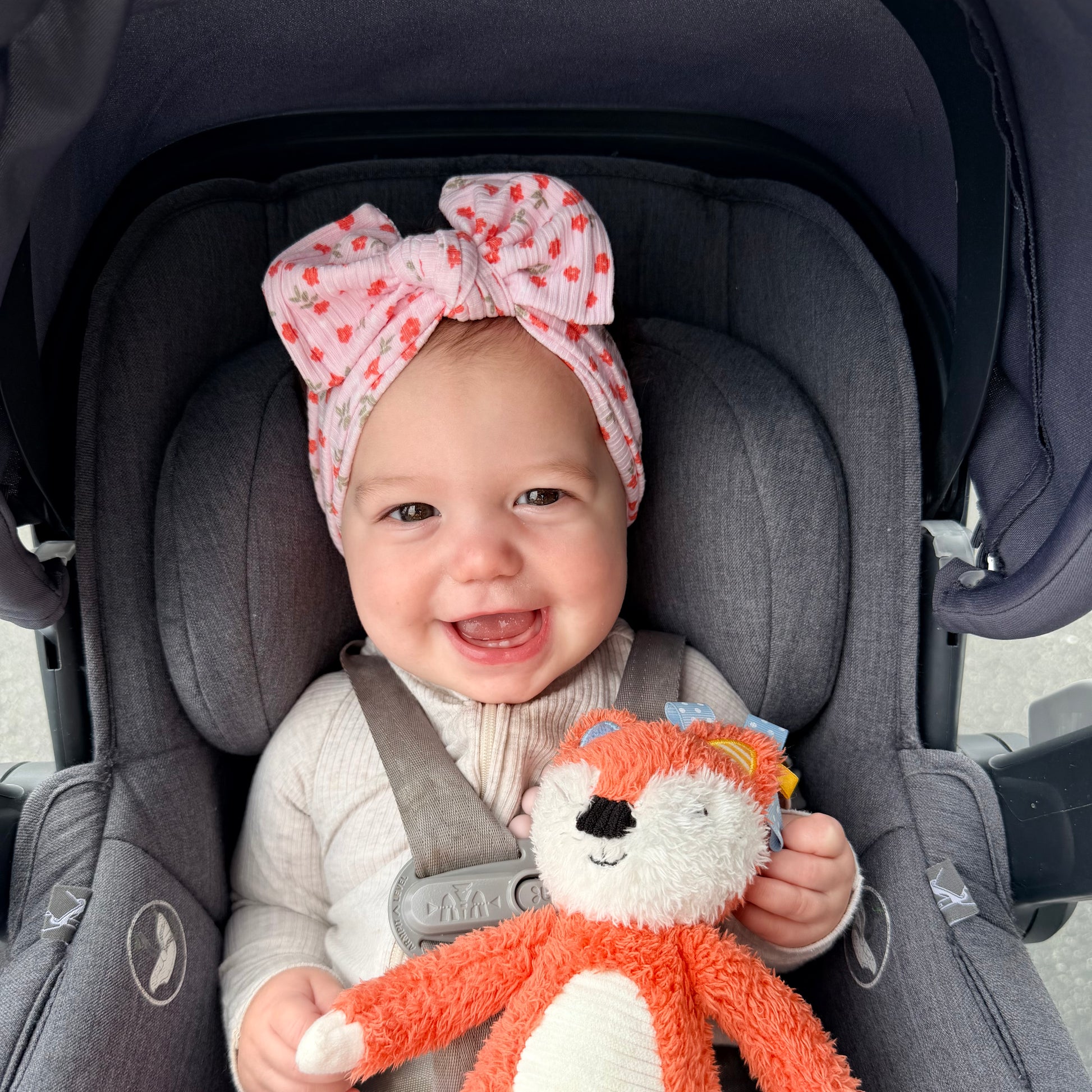 Baby in a car seat wearing a light pink floral ribbed top knot headband and holding an orange plush toy