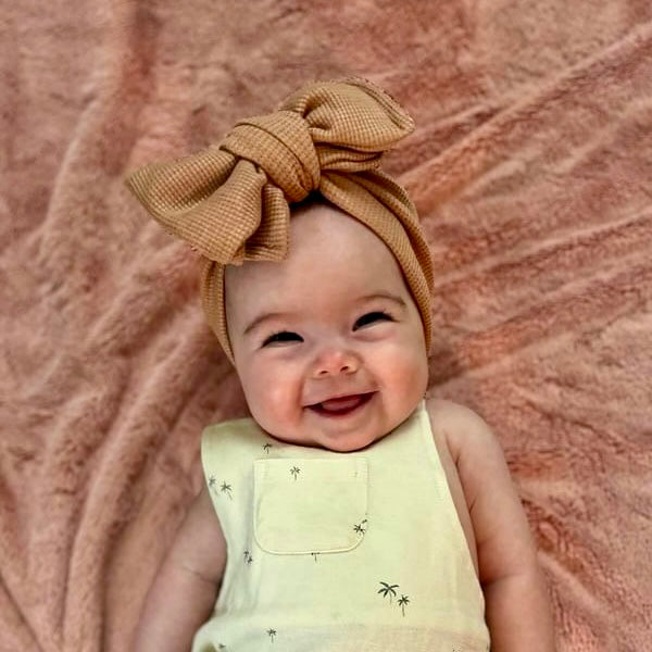 Baby wearing a yellow outfit and mocha brown waffle headband against a textured pink background